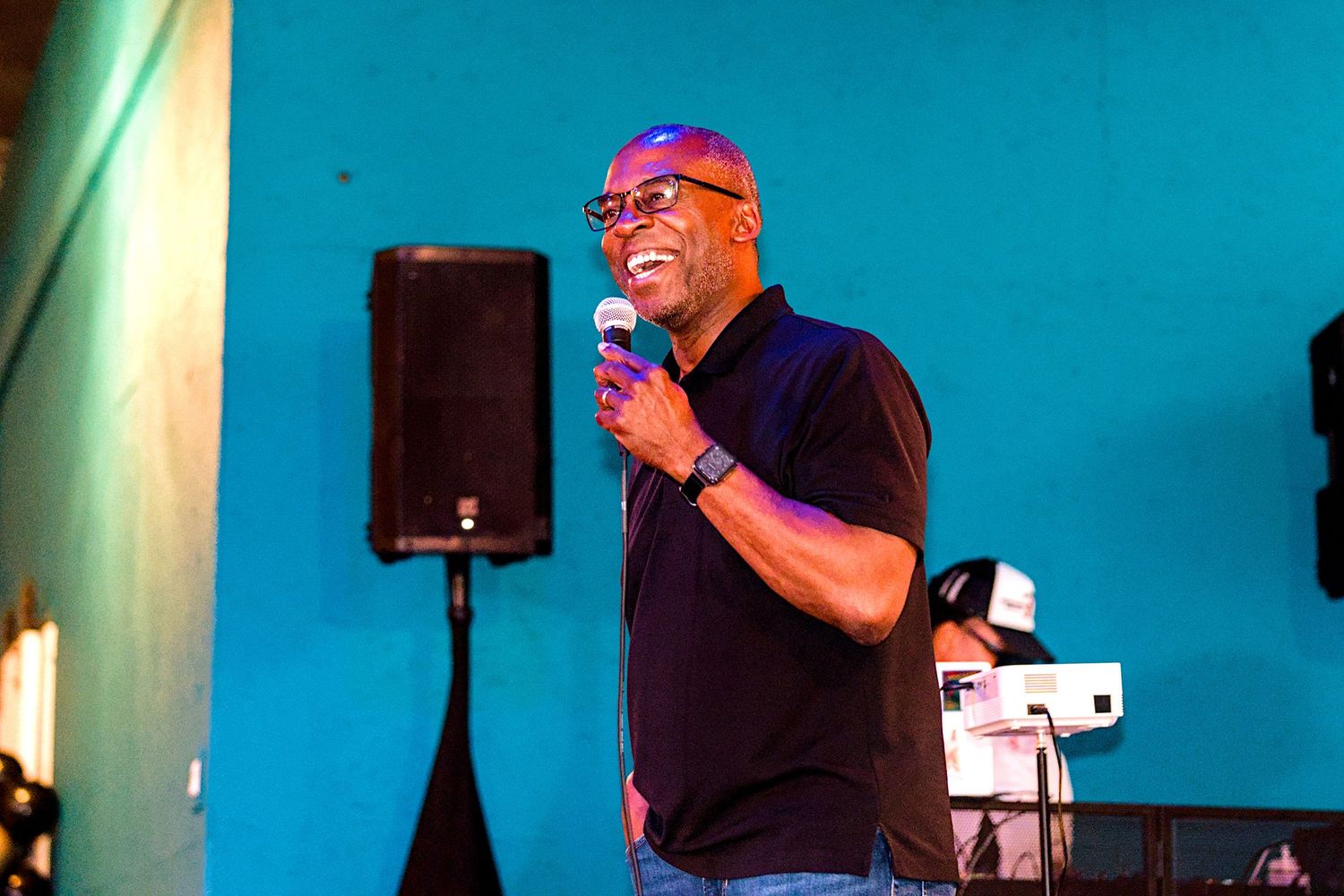 A man smiles while speaking into a microphone on stage in front of a teal wall at an indoor event.
