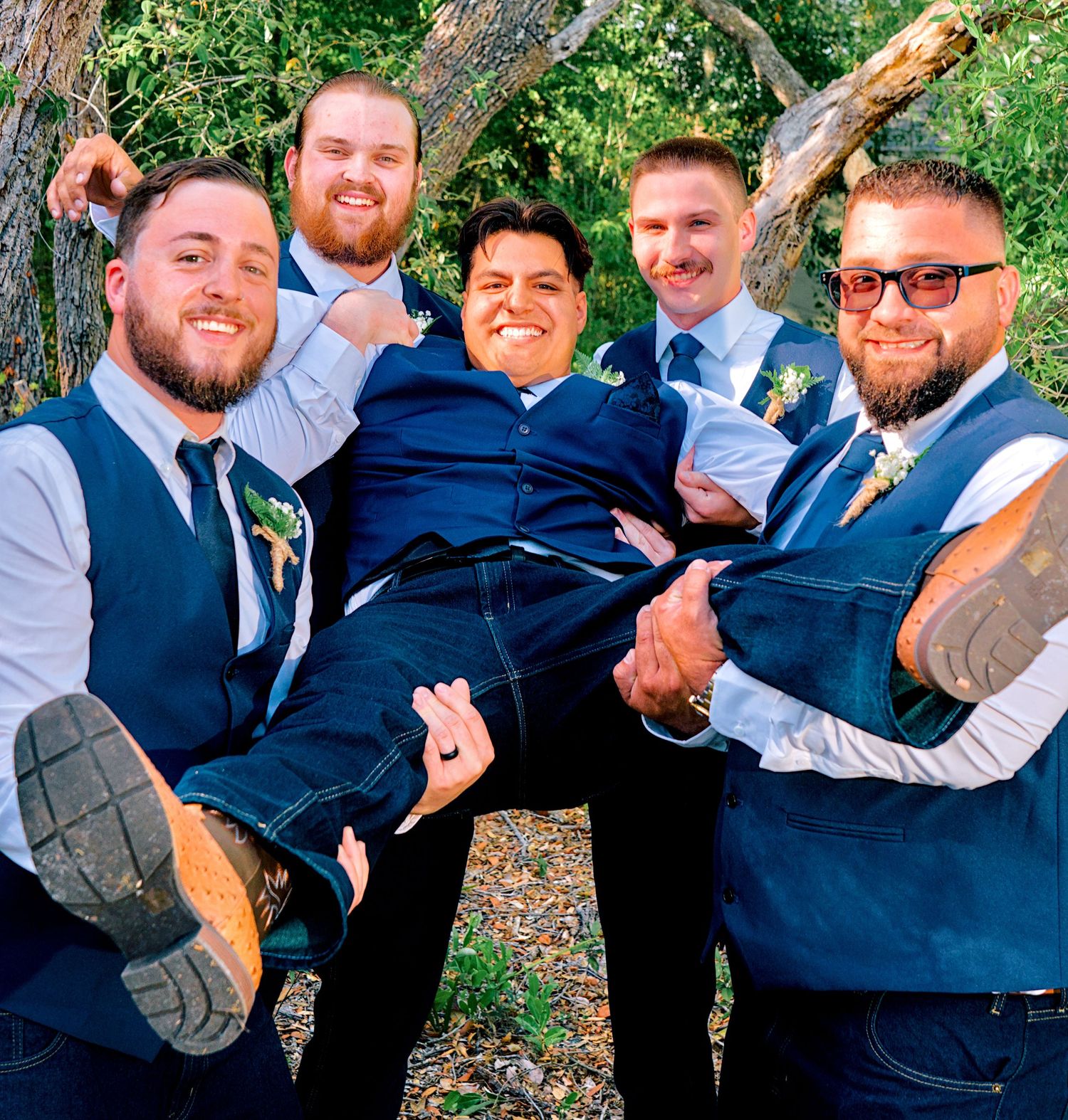 Groom being lifted by four groomsmen in navy vests and jeans in a wooded outdoor setting.