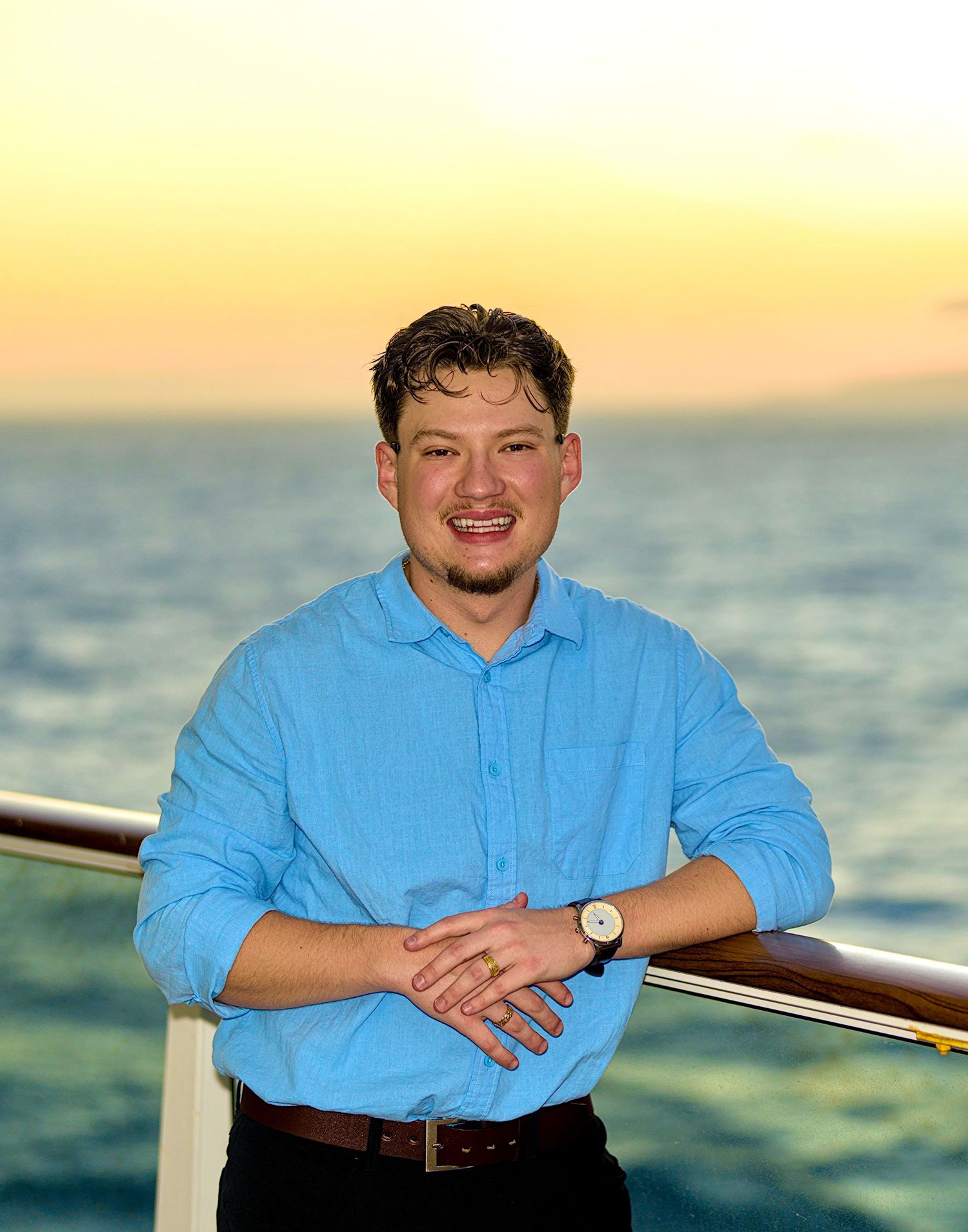 Smiling young man in light blue shirt leaning on railing of cruise ship at sunset over ocean.