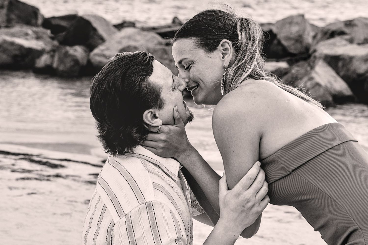 Black and white photo of a romantic couple embracing and smiling at each other on a rocky beach.