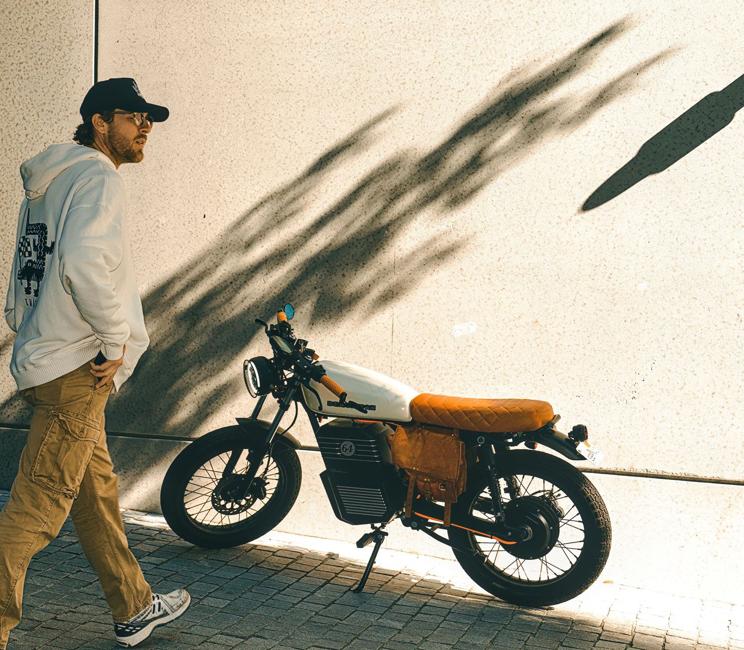 Man in hoodie and cap walking beside a vintage custom scrambler motorcycle against a sunlit concrete wall.