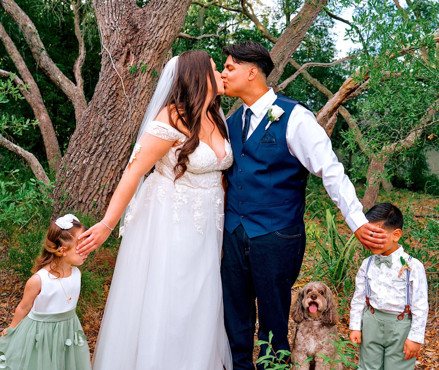 Bride and groom kissing at outdoor wedding with flower girl, ring bearer, and dog beside large tree.