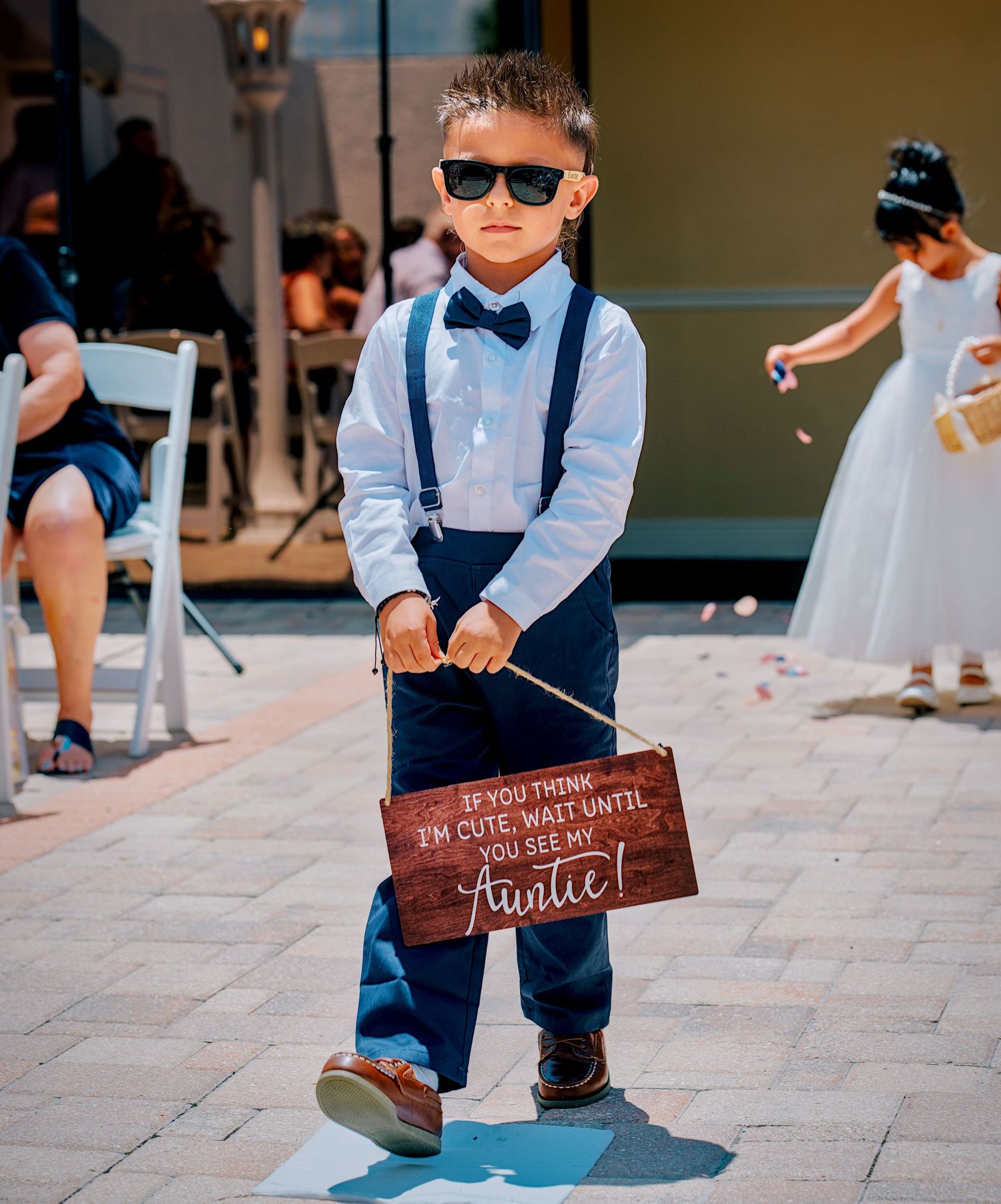 Adorable ring bearer in sunglasses and bow tie holds a wooden sign at an outdoor wedding ceremony.