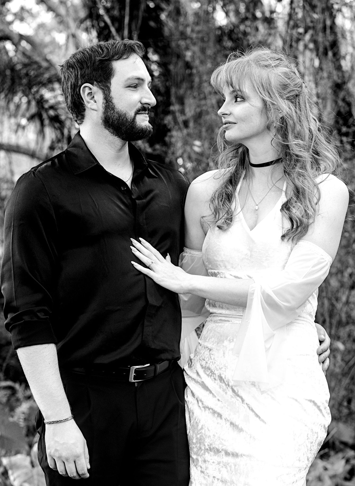 Black and white photo of a couple in formal attire sharing a romantic moment outdoors among lush greenery.