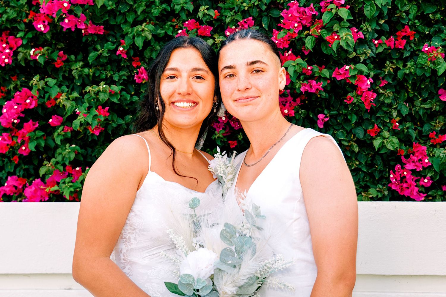 Two brides in white dresses smile together holding a bouquet in front of a vibrant pink bougainvillea wall.