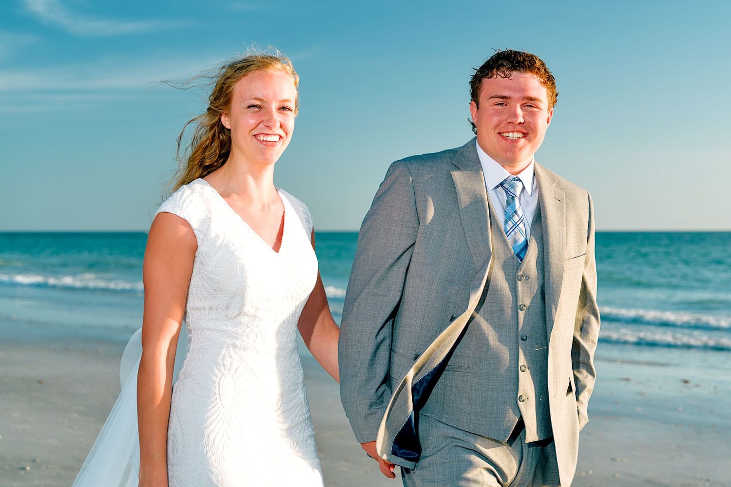 Happy couple in wedding attire smiling on a sunny beach, bride in white gown and groom in gray suit.