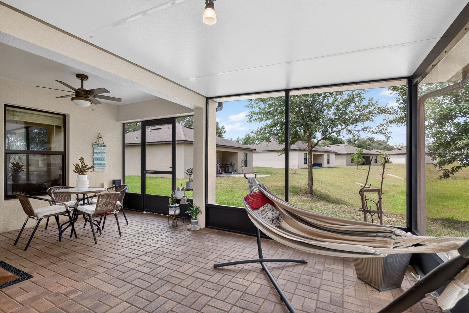 Screened patio with brick flooring, hammock, ceiling fan, outdoor seating, and lush backyard views.
