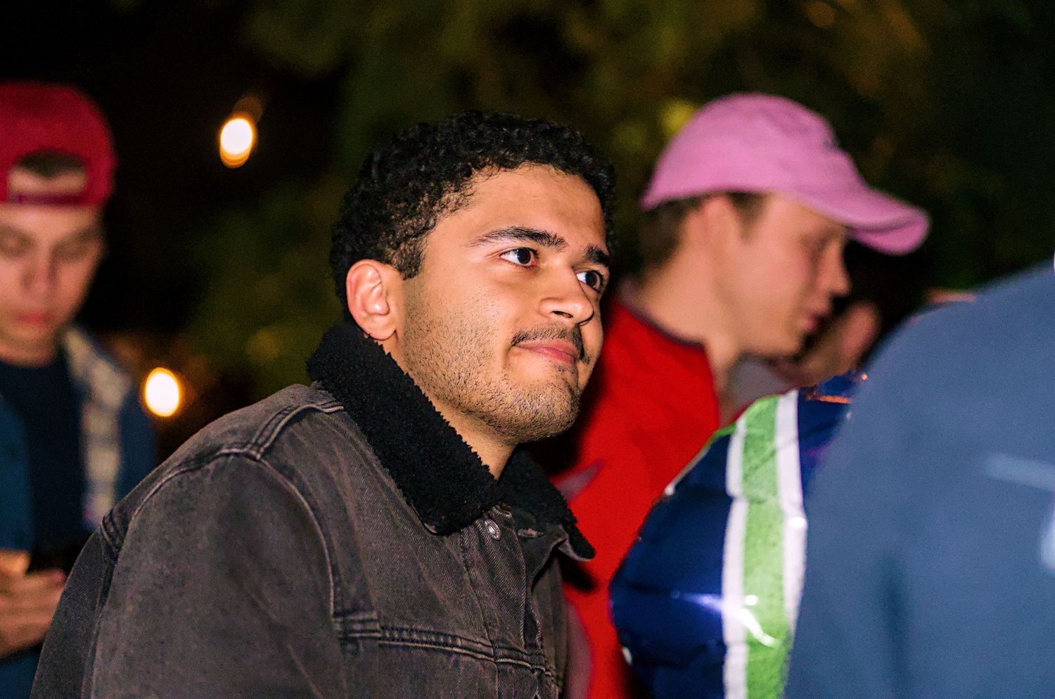 A person in a denim jacket with black collar stands in a dimly lit outdoor setting.