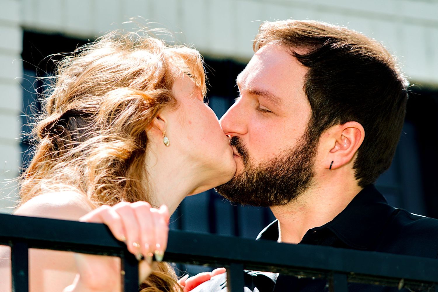 A couple sharing a romantic kiss outdoors near a railing on a sunny day.