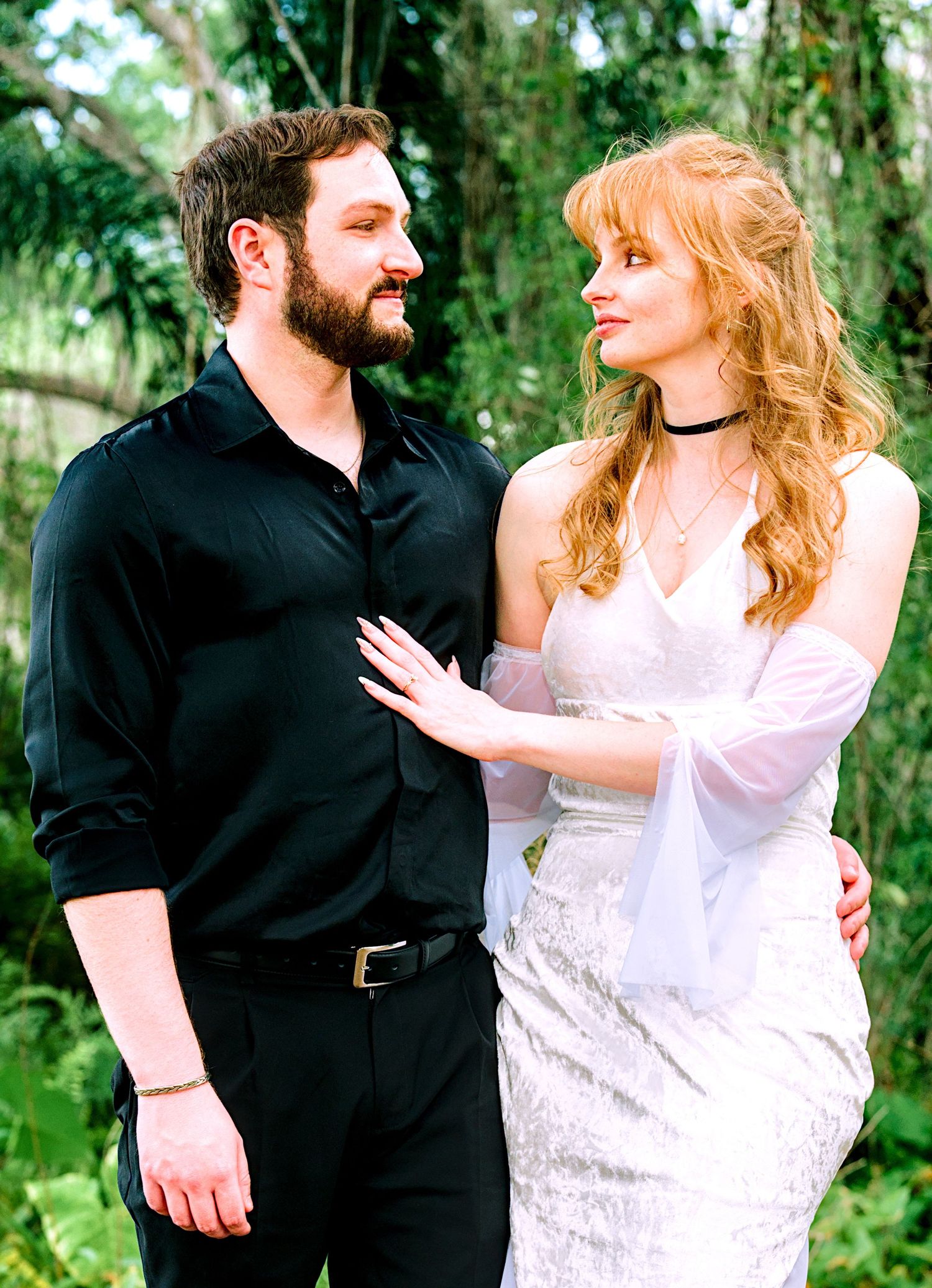 Couple in lush green garden, woman in white lace dress and man in black shirt smiling.