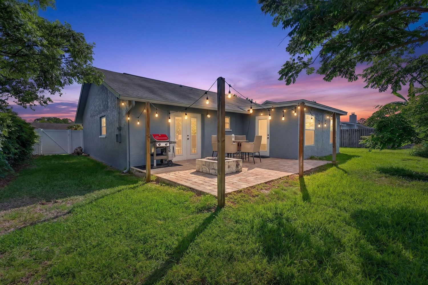 Gray house with string lights over patio at dusk, lush green lawn and trees in warm twilight glow.