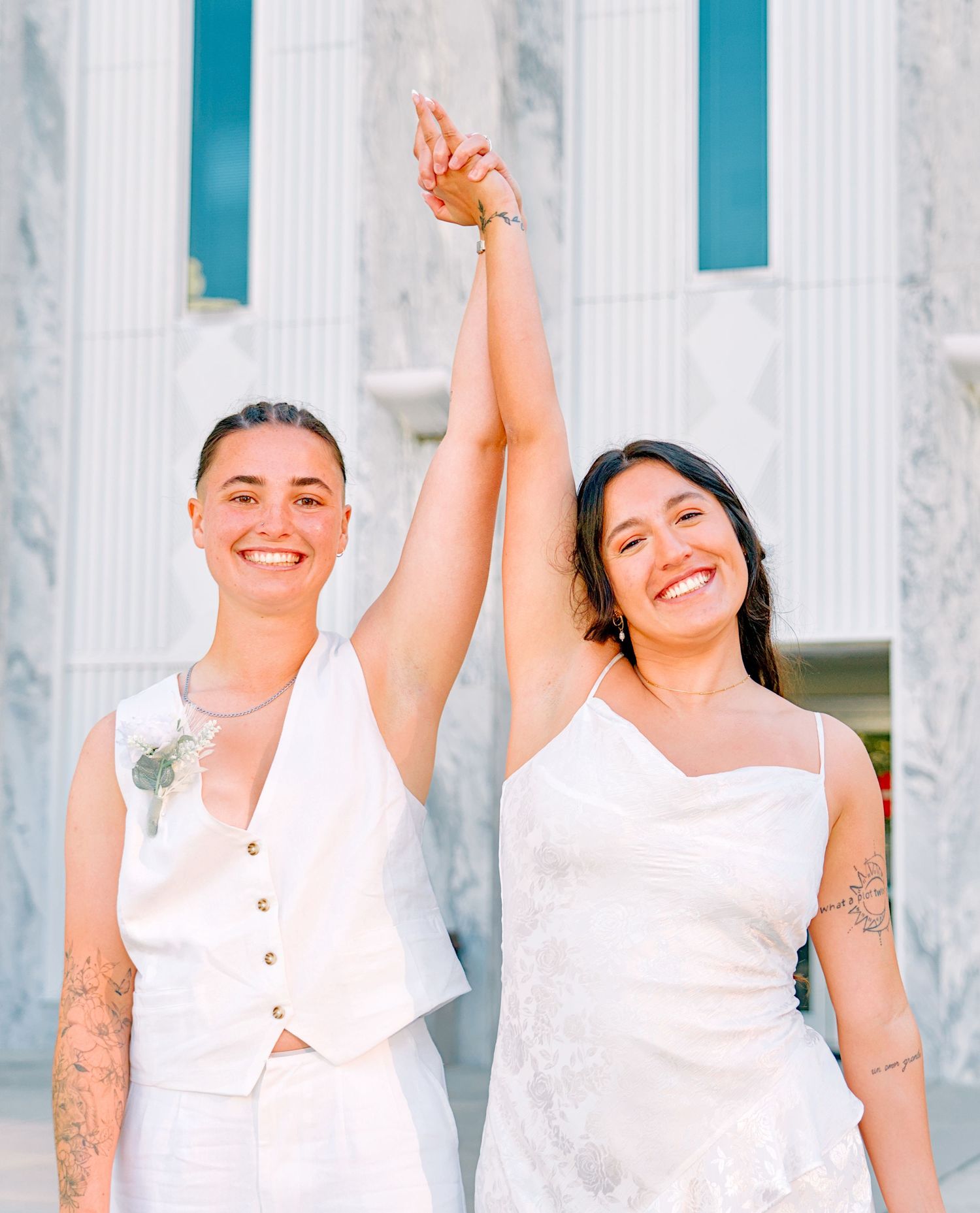 Two brides in white celebrate their wedding day, raising hands together and smiling outdoors.
