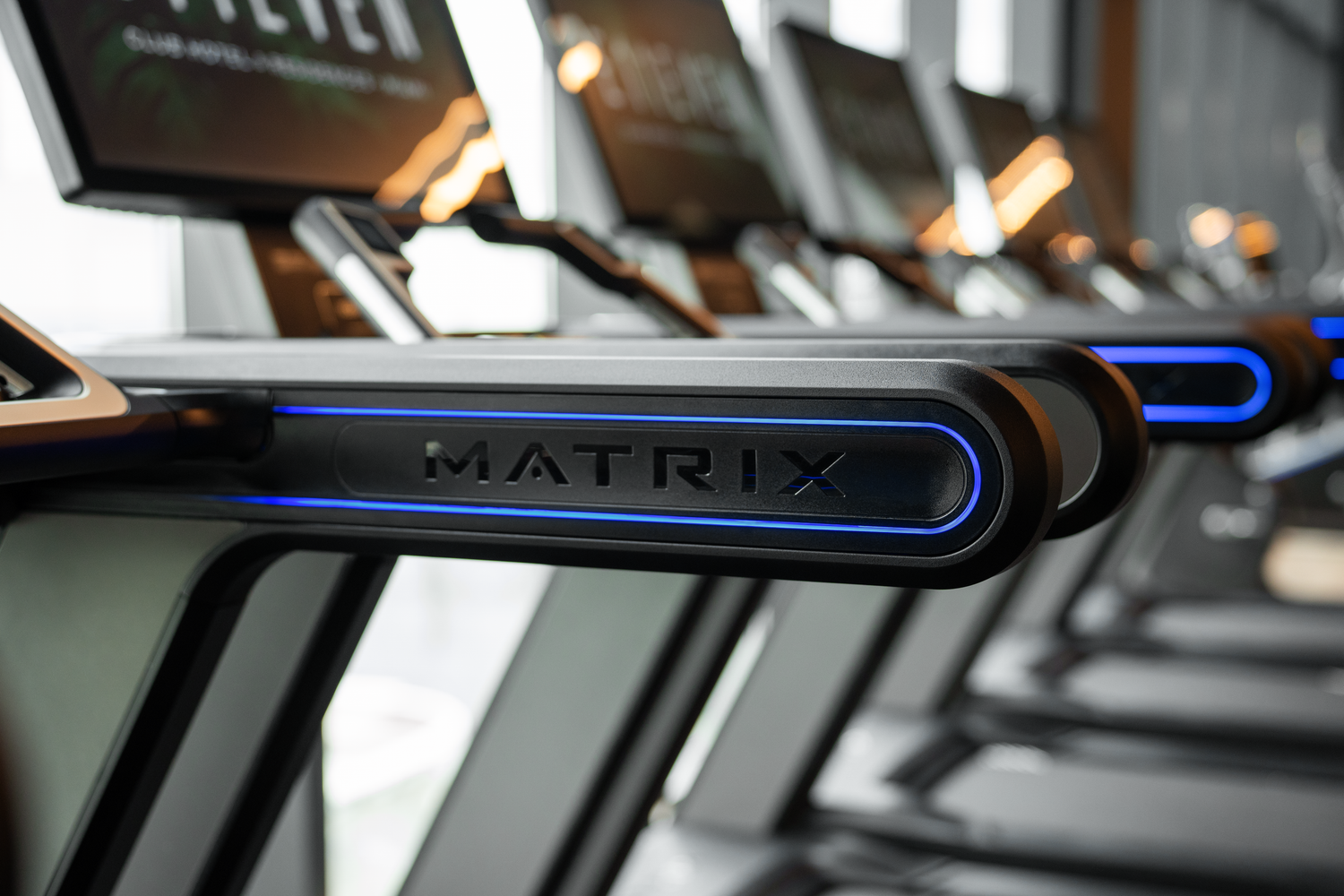 Row of Matrix treadmills in a modern gym with blue LED-lit handrails and digital display consoles.