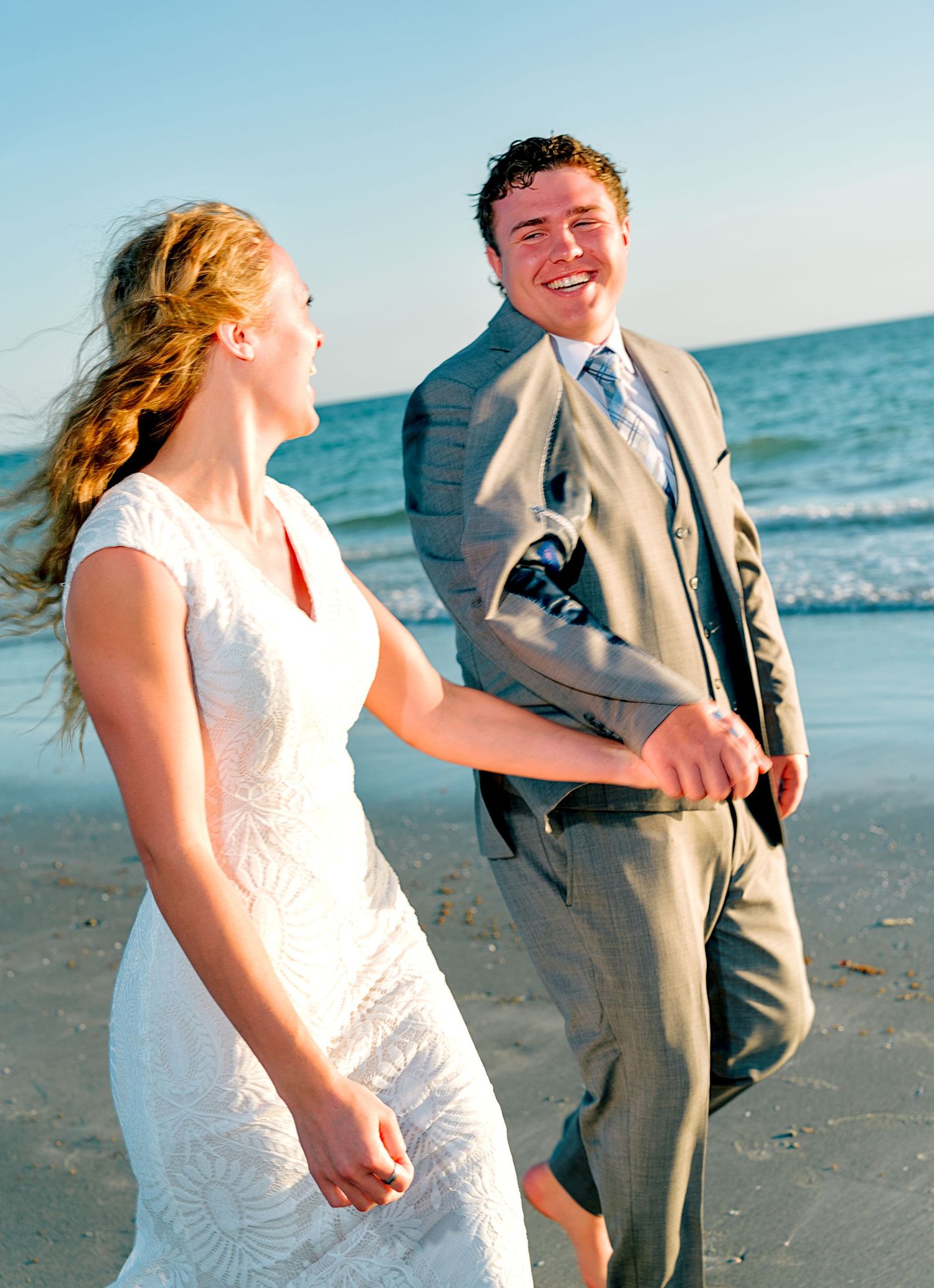 Bride and groom laughing and holding hands on a sunny beach, with ocean waves in the background.