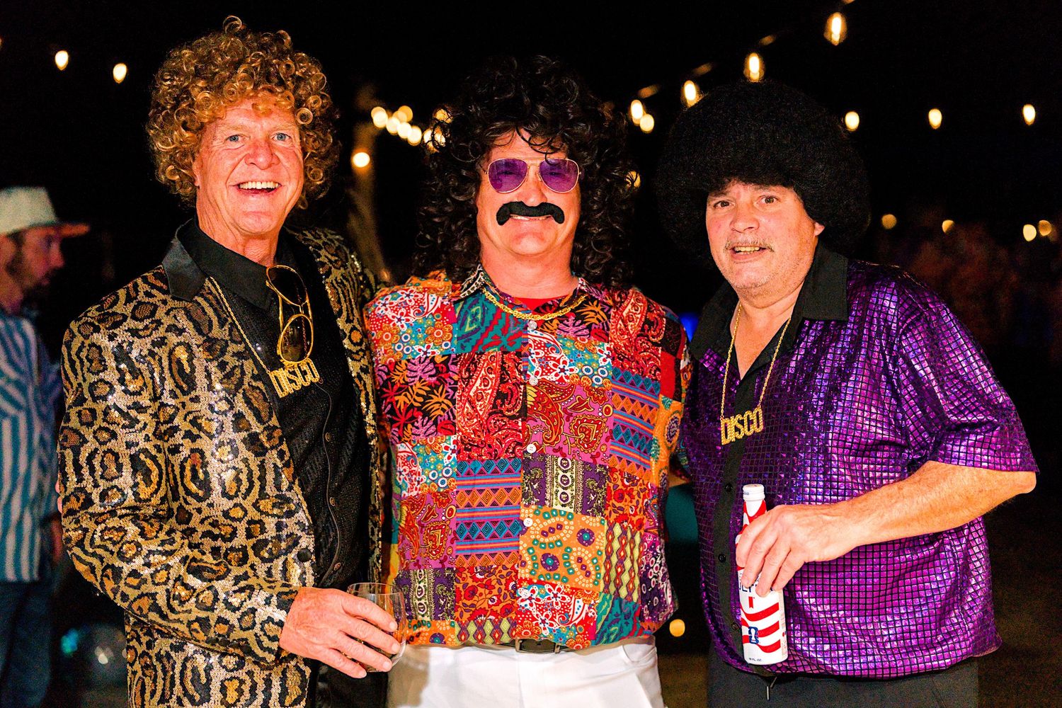 Three men in 1970s disco costumes with afro wigs and sequined shirts posing together at an outdoor themed party.