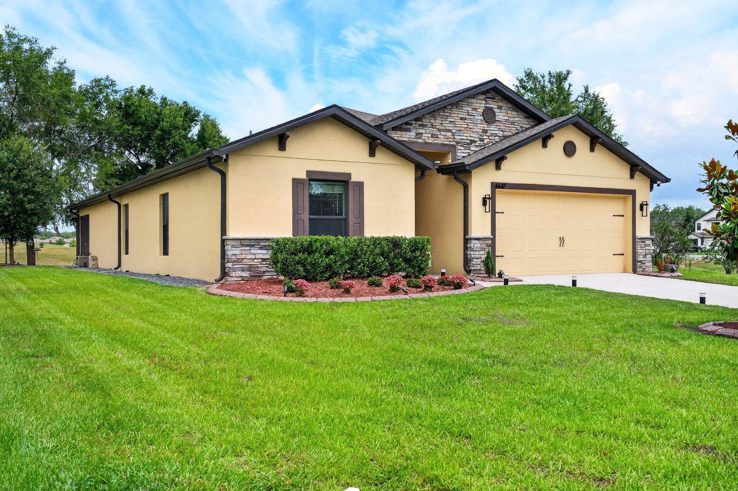 Single-story home with tan stucco exterior, stone accents, attached garage, and lush green lawn under a partly cloudy sky.