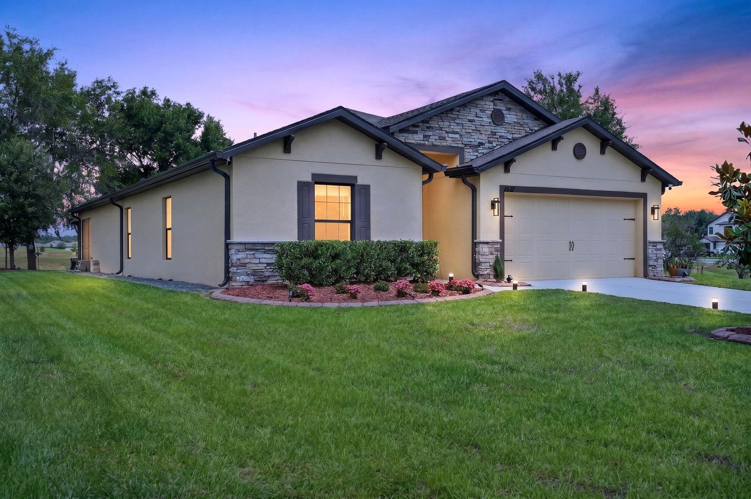 Single-story stucco home with stone accents, two-car garage, lush green lawn, and colorful sunset sky in background.