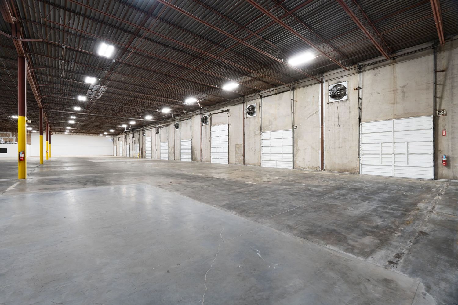 Large empty industrial warehouse interior with concrete floors, white walls, and overhead lighting stretching into the distance.