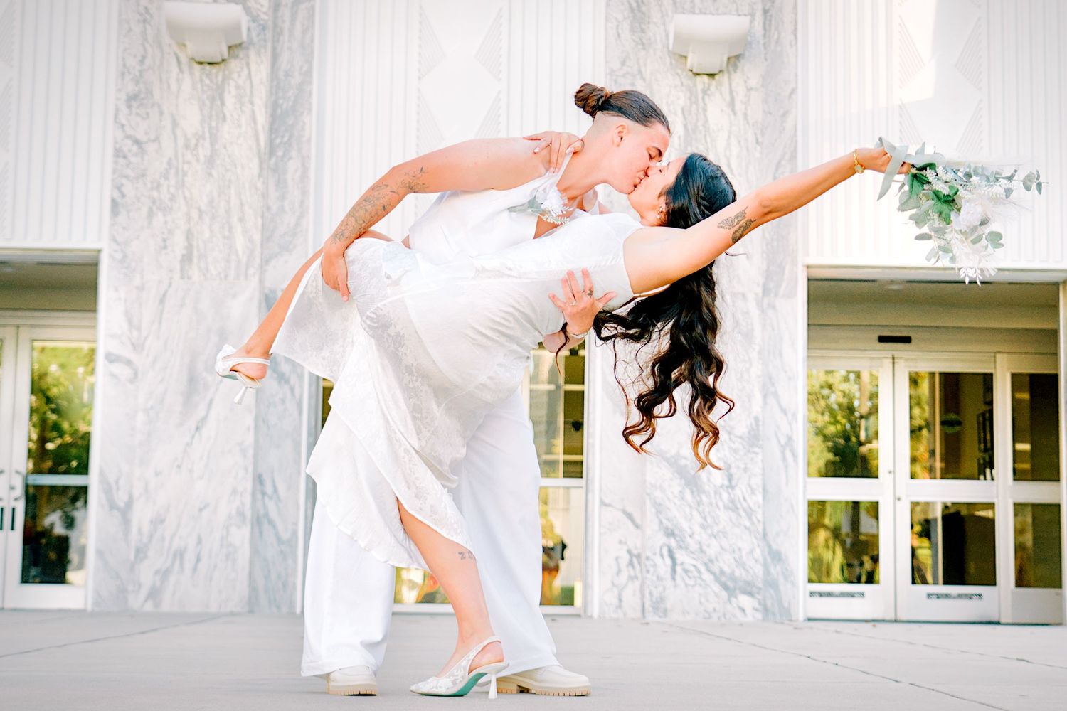 Couple sharing a romantic dip kiss in wedding attire outside elegant marble columns building.