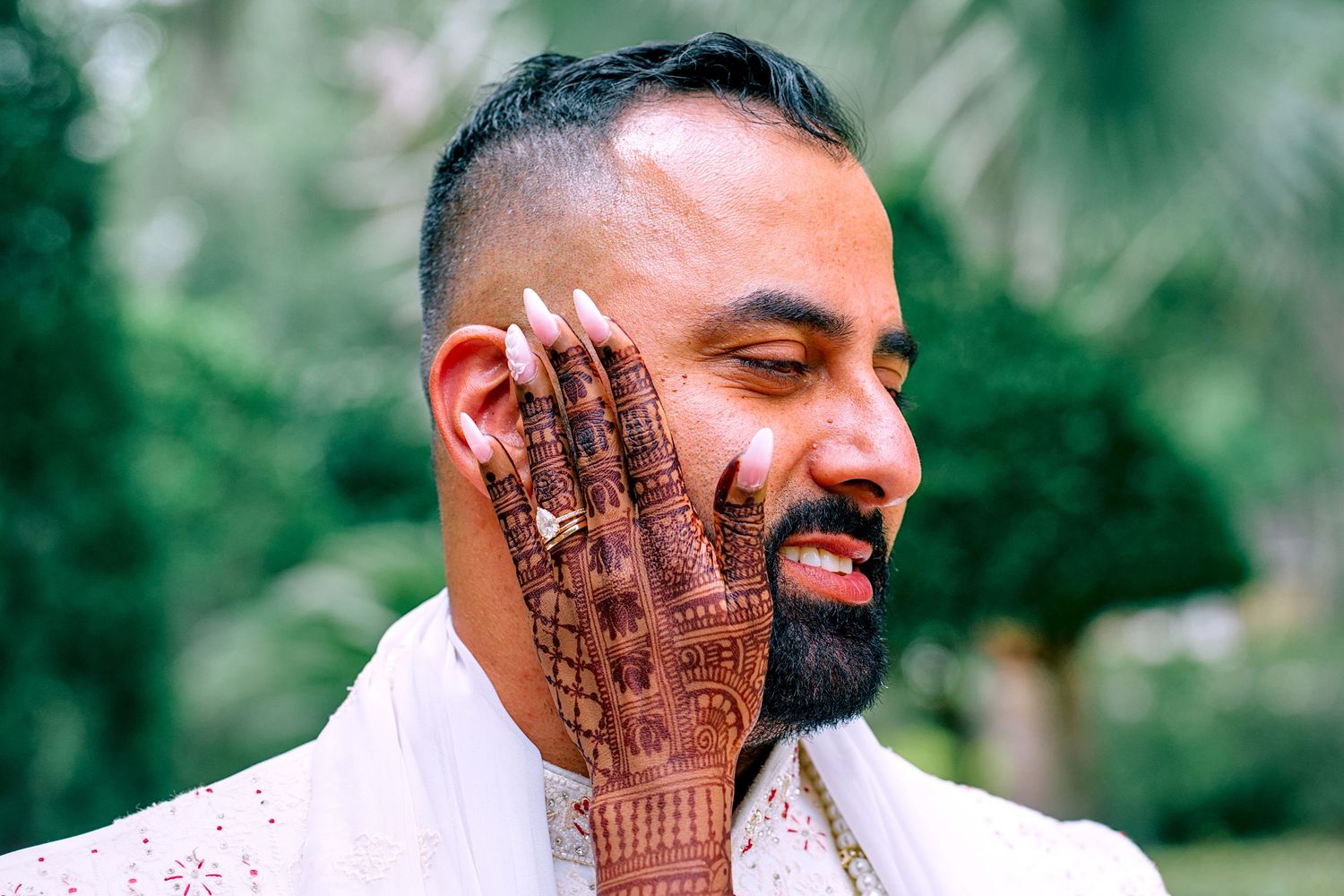 Indian groom with henna-adorned hand touching his face outdoors.