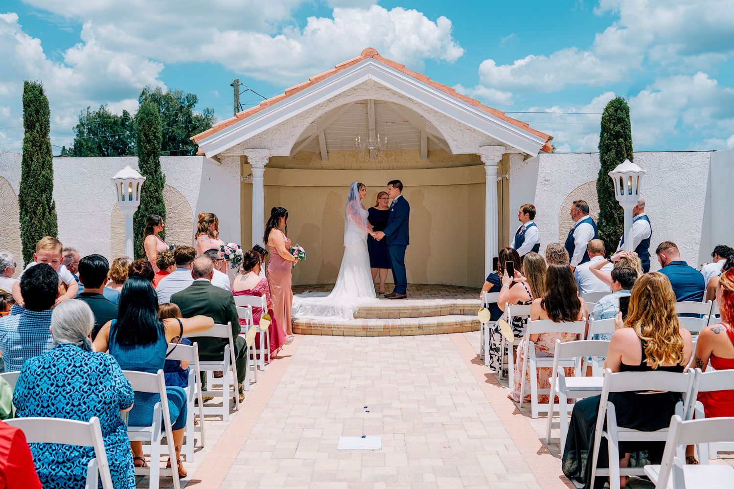 Outdoor wedding ceremony at elegant white chapel with bride and groom at altar, surrounded by seated guests on white chairs.