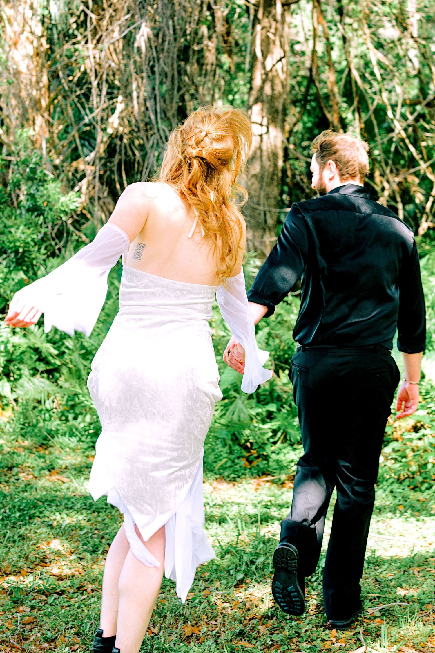 Bride in white dress and groom in black walking hand in hand through lush green woods.