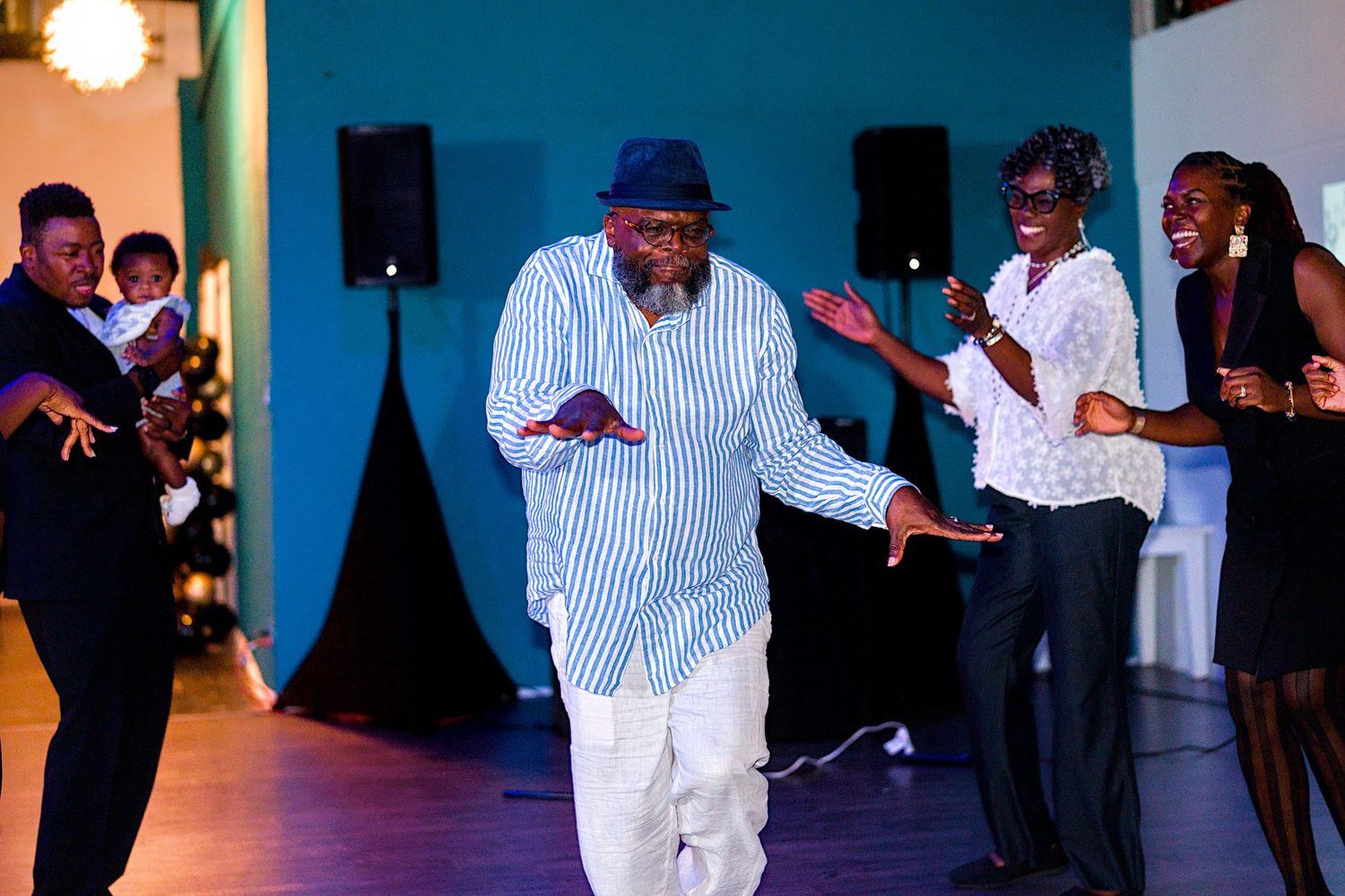 A man in a striped blue shirt dances joyfully on a dance floor surrounded by smiling guests at an event.