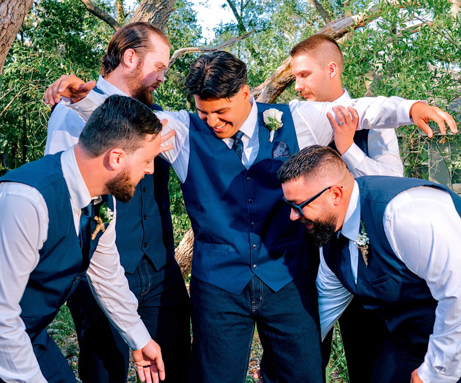 Groom in blue suit surrounded by groomsmen in matching vests celebrating at an outdoor wedding ceremony.