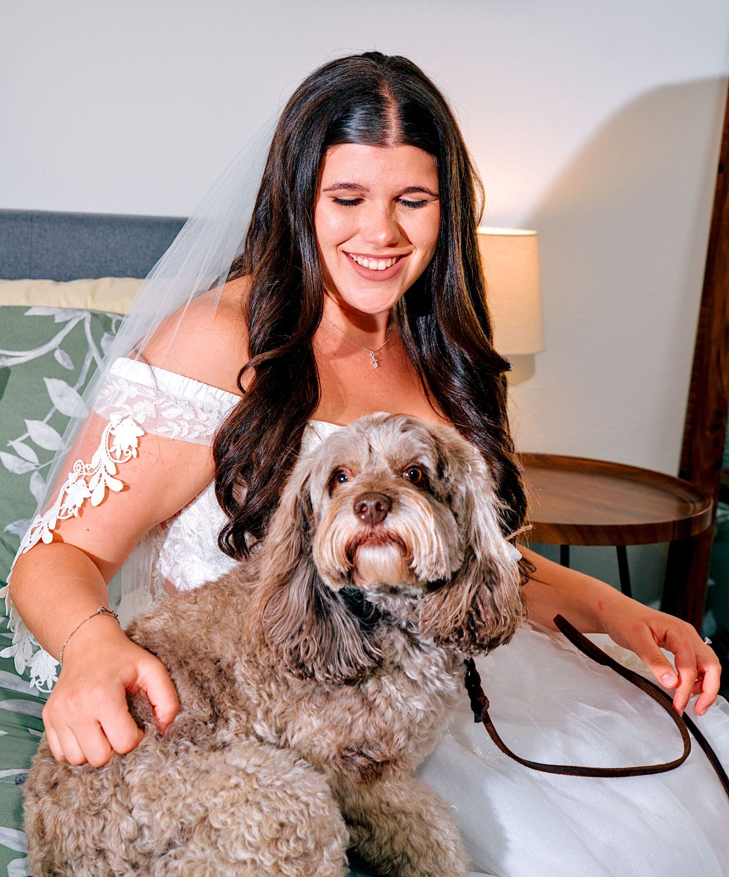 Smiling bride in white off-shoulder gown and veil sits with fluffy gray Cavapoo dog on leash indoors.