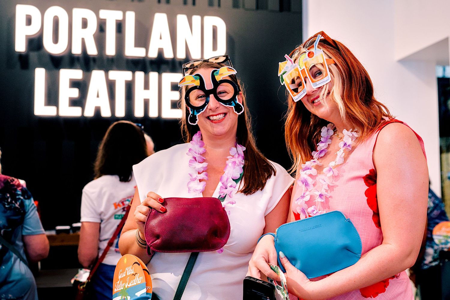 Two women wearing fun glasses and leis smile holding Portland Leather bags at a Portland Leather store event.