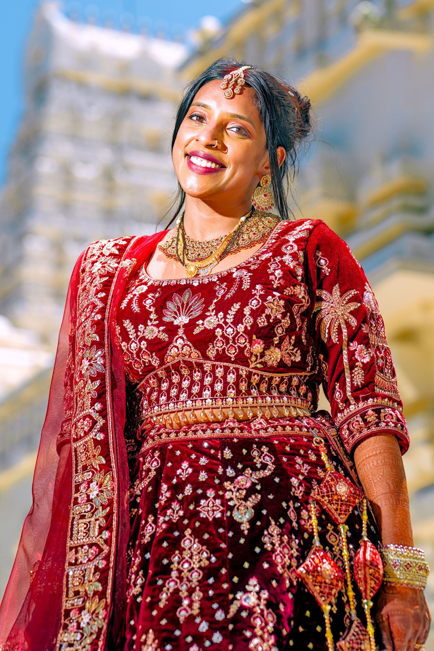 Indian bride in red embroidered lehenga with gold jewelry smiling outdoors.
