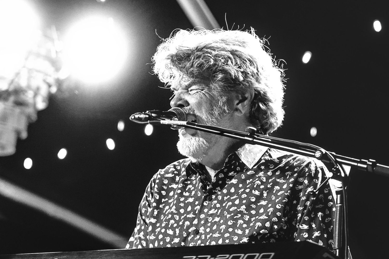 Black and white photo of a musician playing keyboard on stage under bright spotlights at a live concert.
