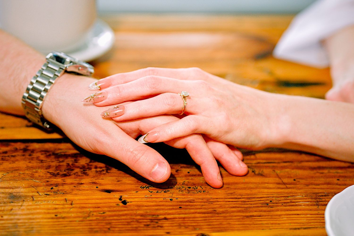 Two hands resting on a wooden table, both wearing gold rings, one with a silver watch.
