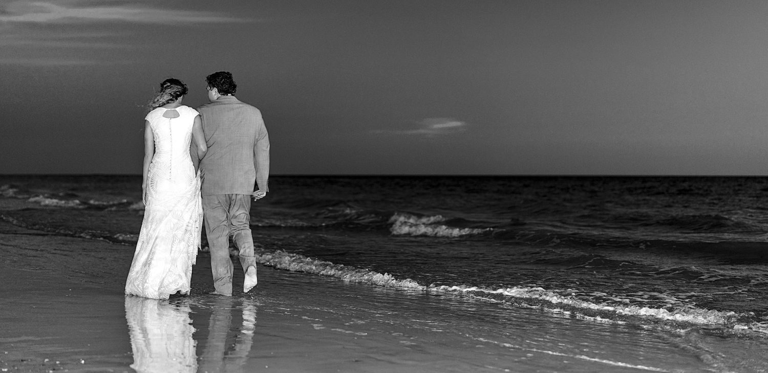 Black and white photo of a bride and groom standing together on a beach shore, gazing at the ocean waves.