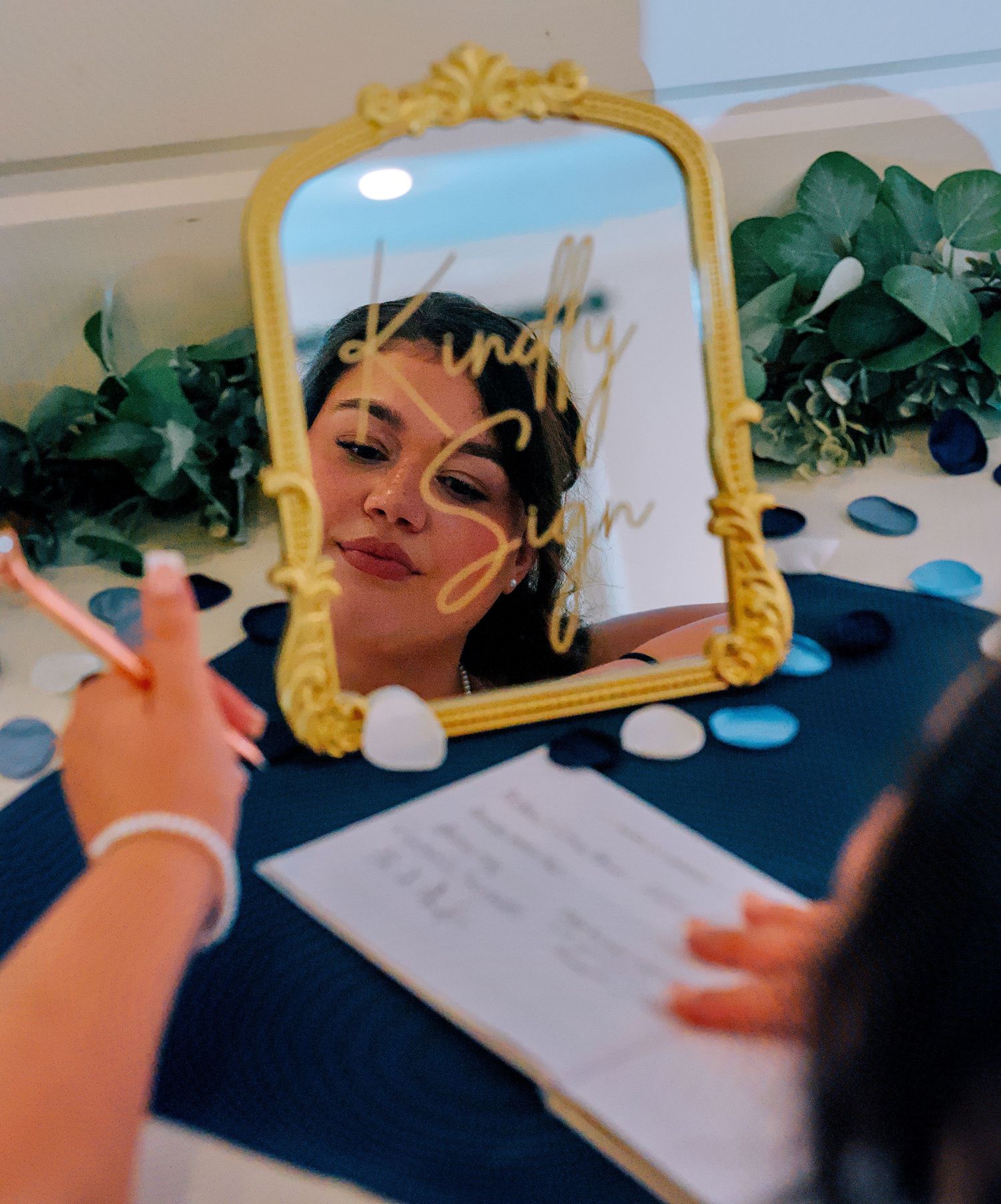 A woman reflects in a gold ornate mirror at a wedding sign-in table decorated with greenery and colorful stones.