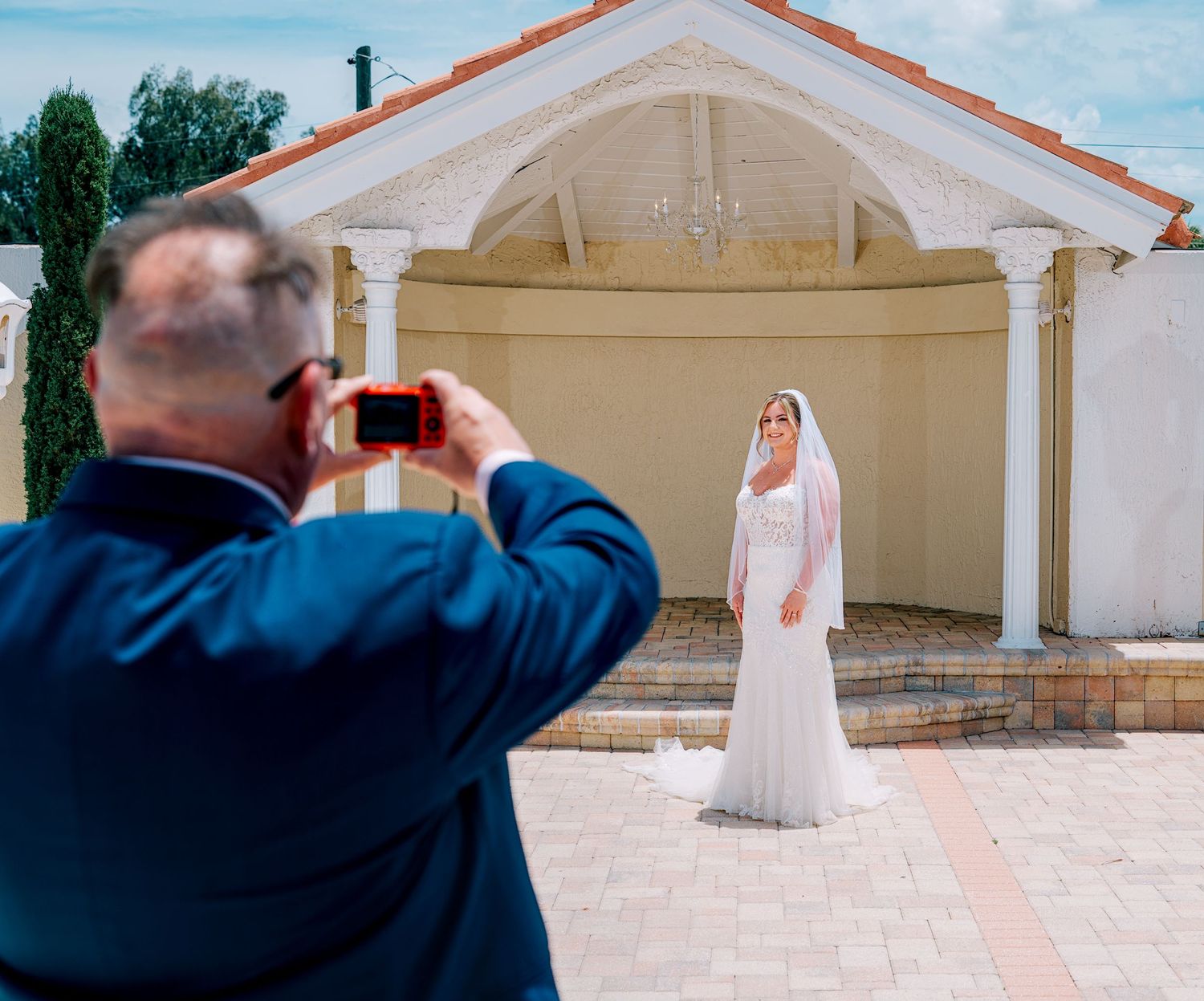 Man in blue suit photographs bride in white gown standing before elegant white gazebo on wedding day.