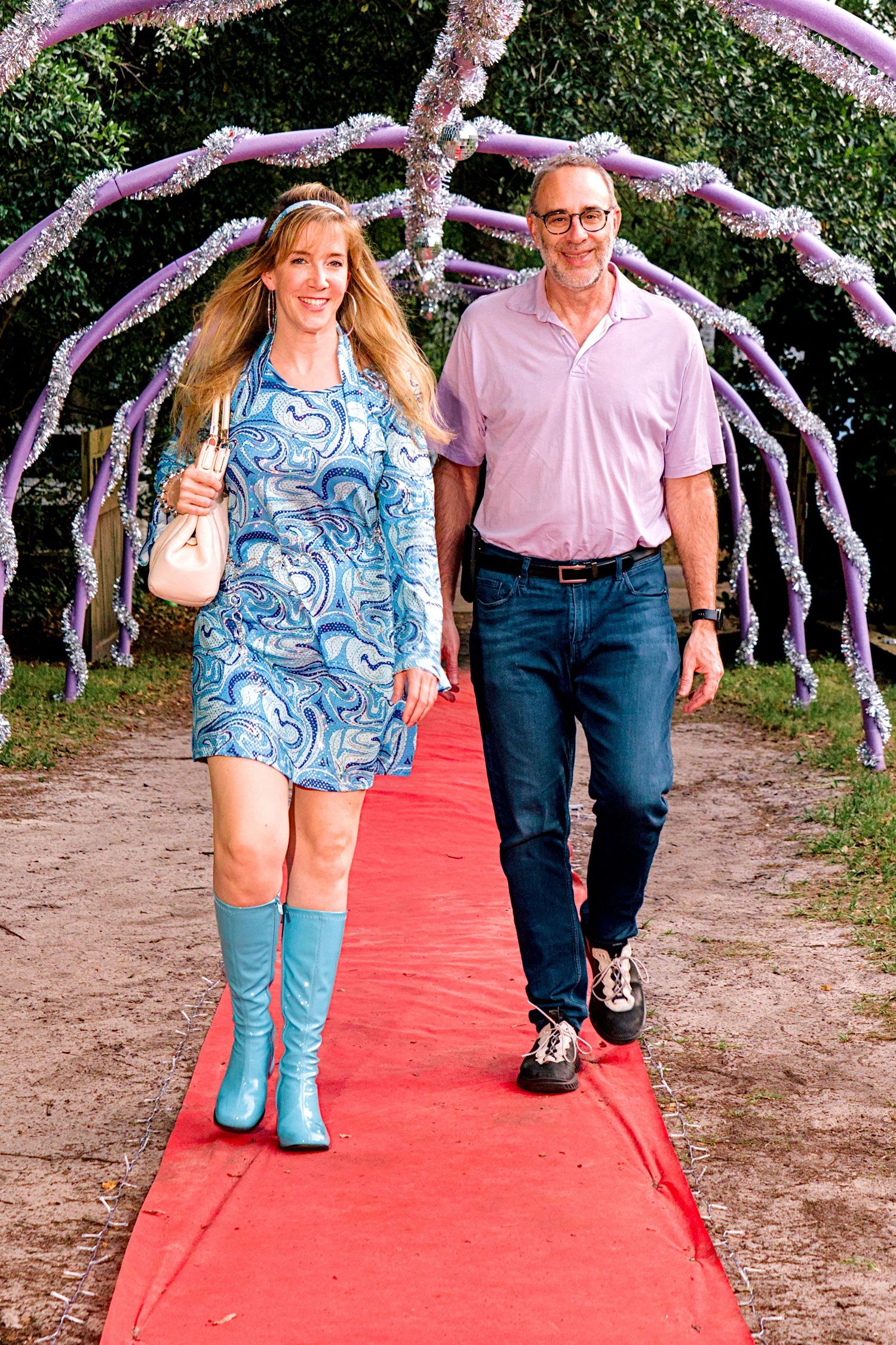 Couple walking down red carpet under purple floral arch at outdoor event.