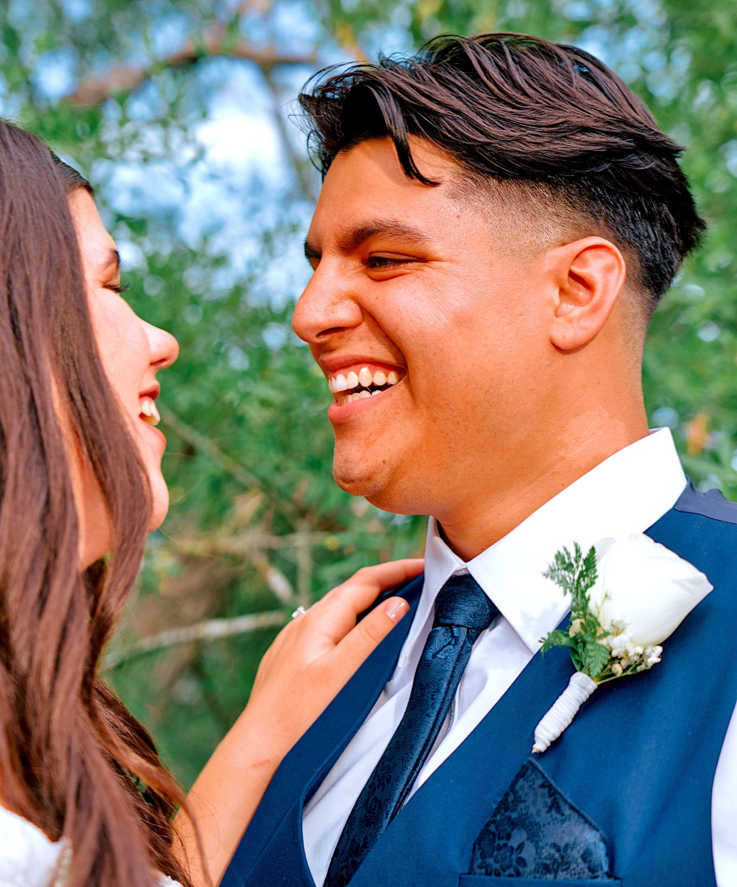Groom in navy suit laughs joyfully as bride adjusts his tie outdoors among green trees.