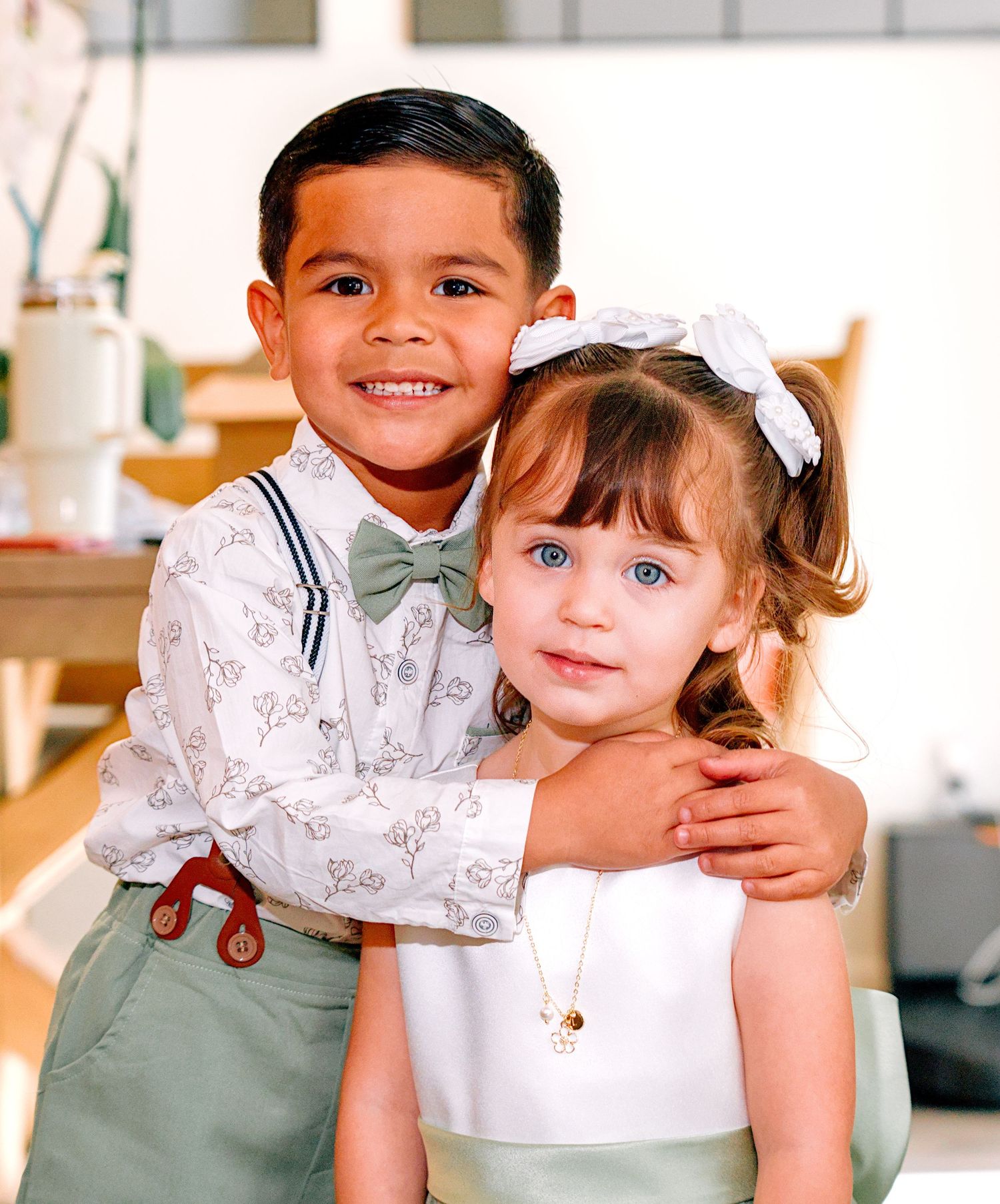 Two young children dressed in formal wedding attire smile together, boy in bow tie and suspenders, girl in white dress.