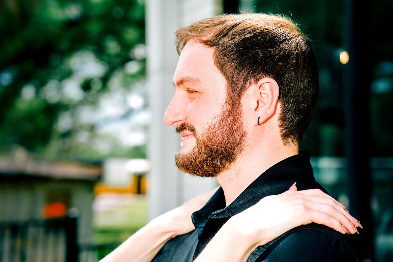 Bearded groom in black tuxedo with bow tie smiling in profile outdoors.