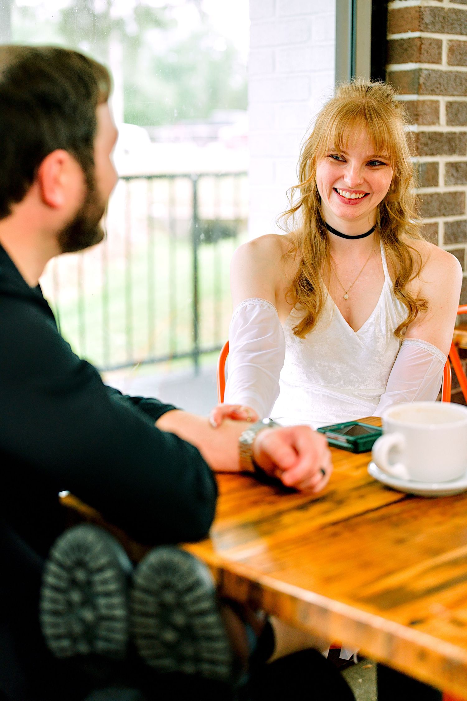 Smiling blonde woman in white dress sitting across from bearded man at wooden cafe table.