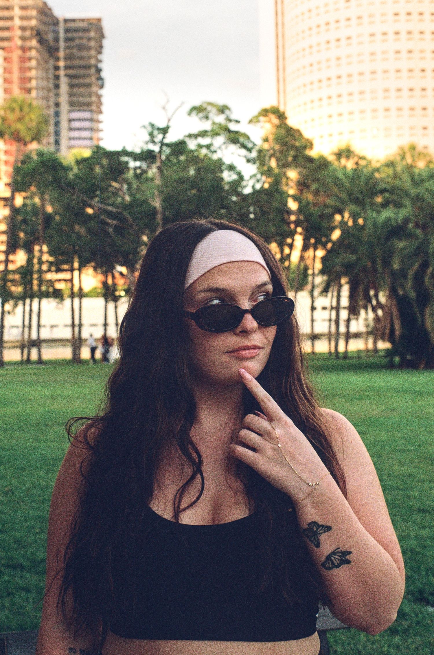 Woman with long dark hair, white headband, sunglasses, and tattoos poses in urban park with city buildings.