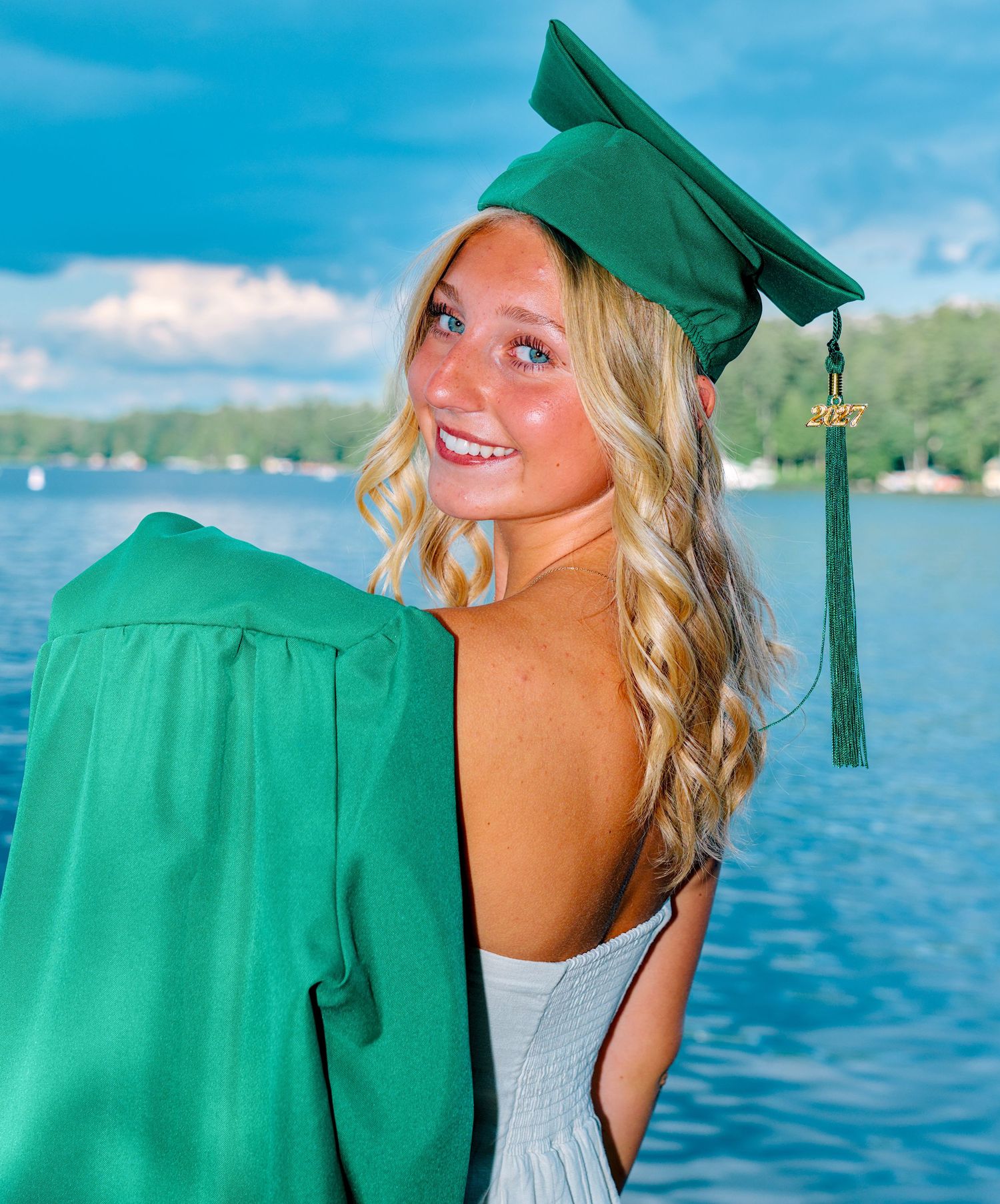 Smiling blonde graduate wearing green cap and gown poses outdoors near a lake on a sunny day.