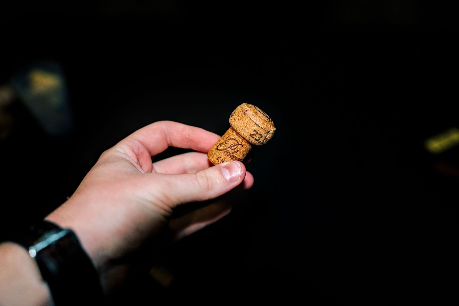 A hand holding a small wine cork against a dark background, wearing a black wristwatch.