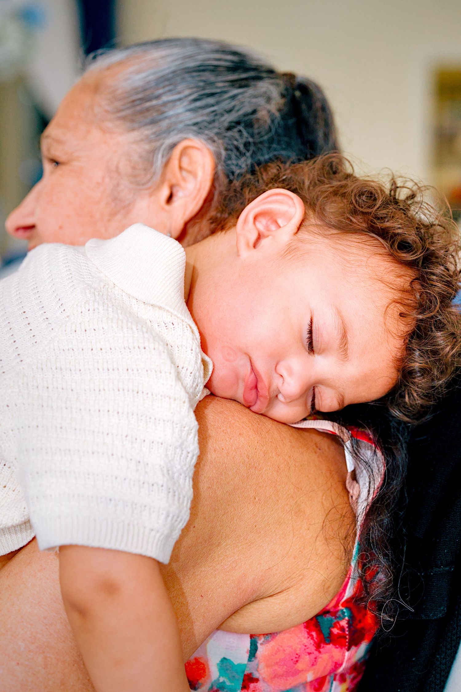 A young child with curly hair sleeps peacefully on an elderly woman's shoulder at a family gathering.