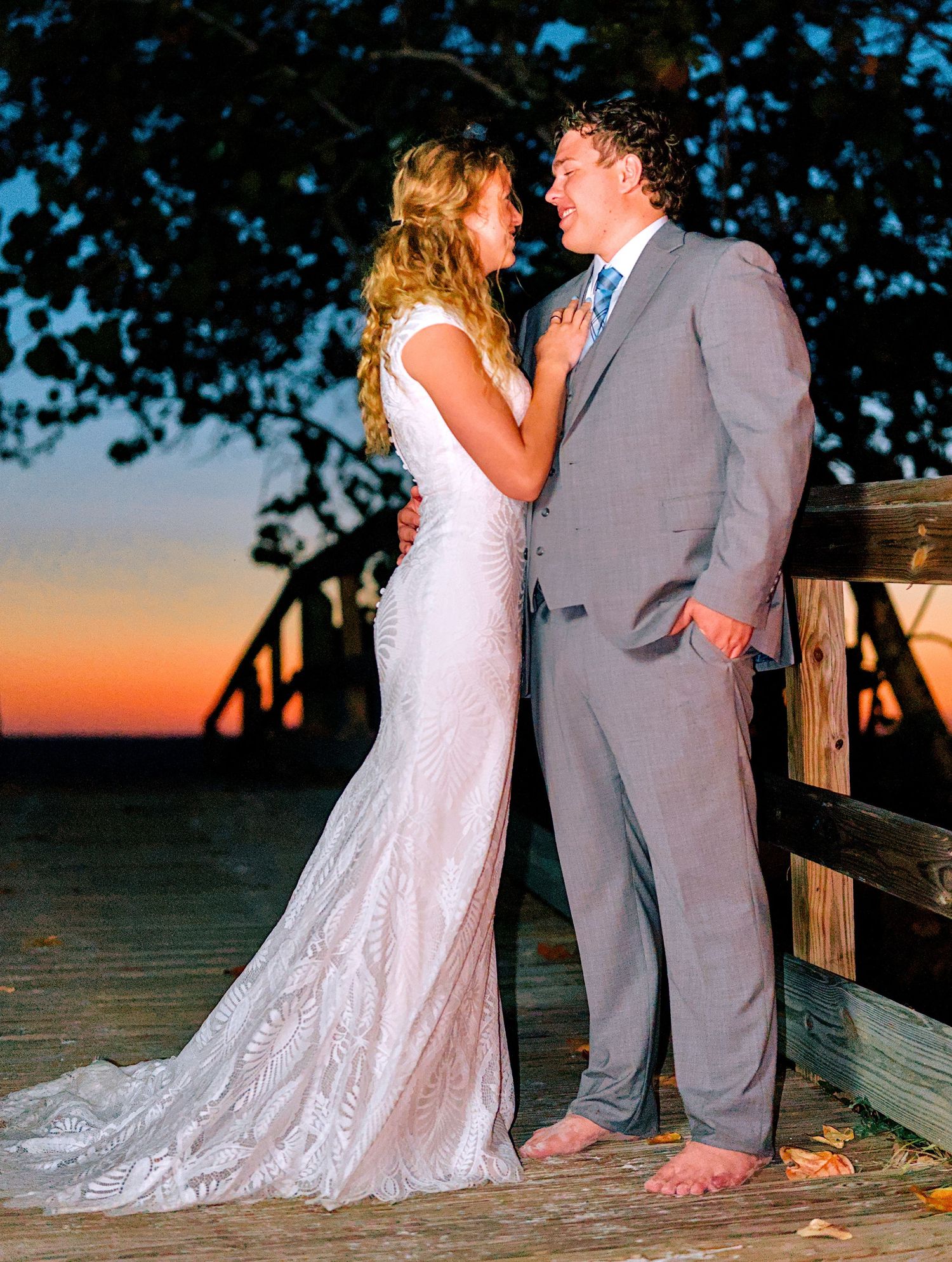 Bride and groom gazing at each other on a wooden deck at sunset, she in a lace gown, he in a gray suit.
