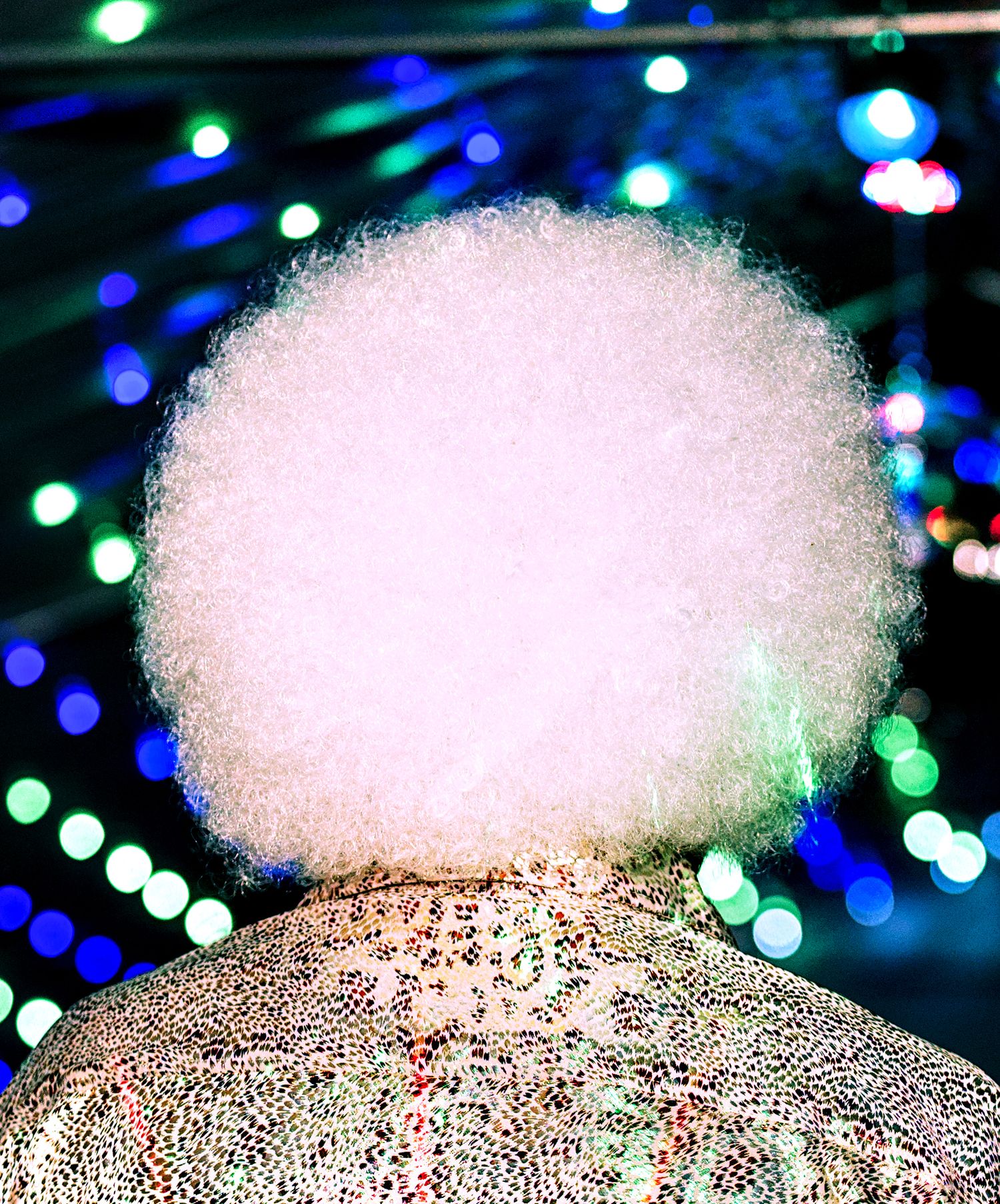 Person wearing white afro wig and sequined jacket posed against colorful bokeh lights background.
