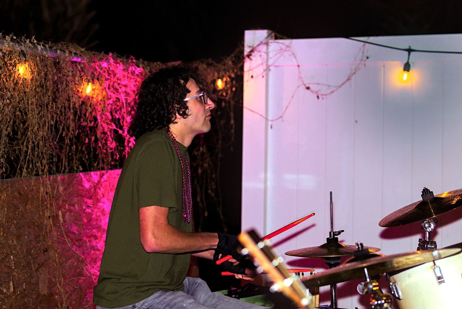 A drummer performs on stage with pink lighting and string lights in the background.