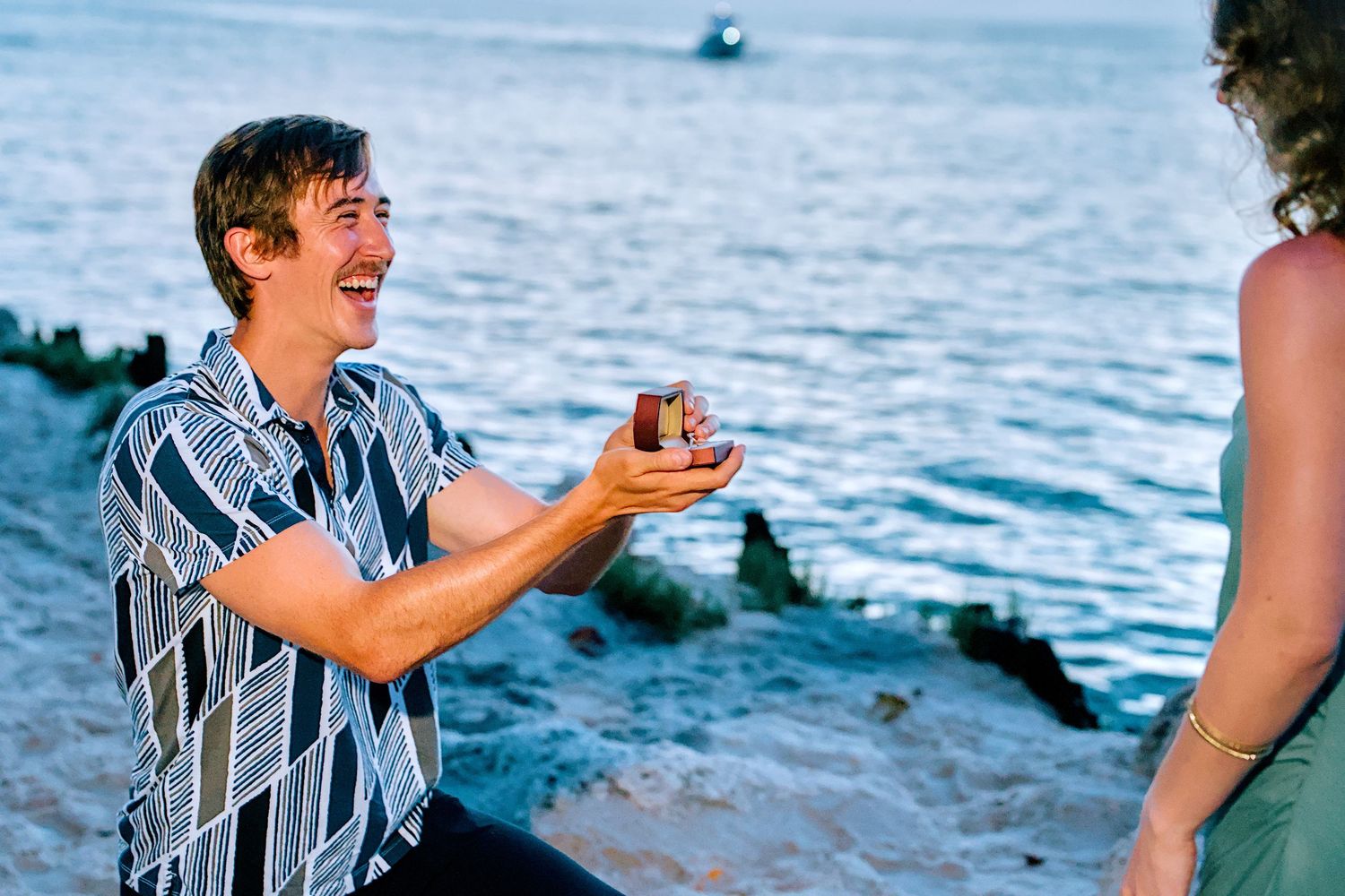 Man kneeling on beach proposing with ring box to woman at sunset near ocean rocks.