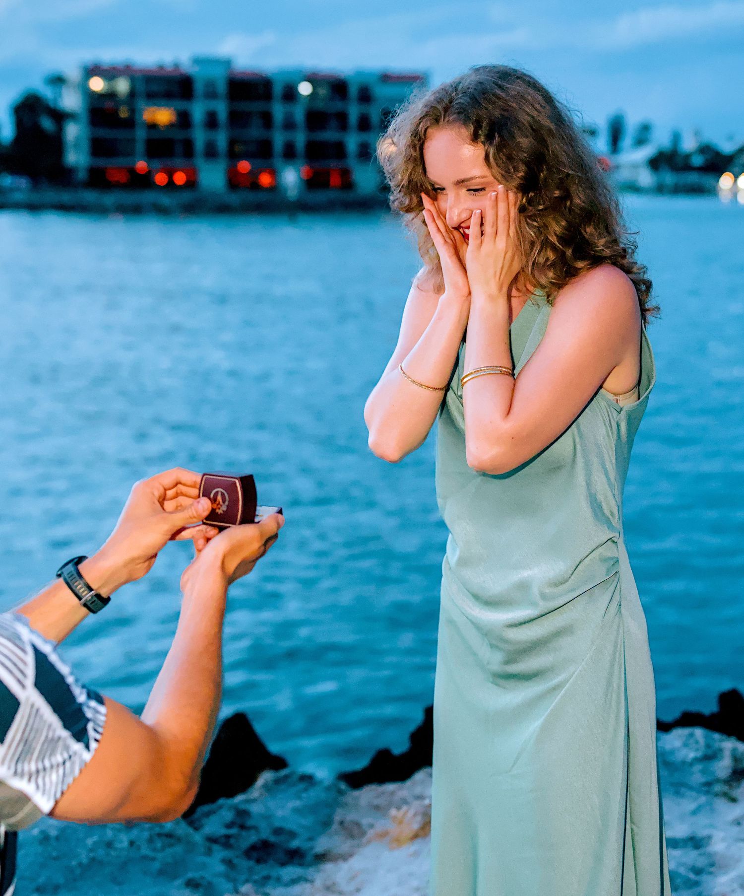 Woman reacting with surprise and joy as someone proposes with a ring box by the waterfront.